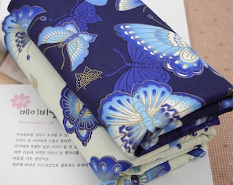 May include: Two folded fabrics with a butterfly pattern. One fabric is navy blue with light blue and gold butterflies. The other is off-white with blue and gold butterflies. The fabrics are stacked on top of each other.