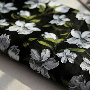 May include: Close-up of black fabric with a white and green floral pattern. The fabric has a textured, woven look.