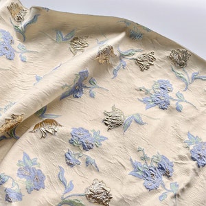 May include: A close-up of a cream-colored fabric with a floral pattern. The flowers are blue and gold, and the fabric has a textured surface.