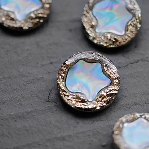 Puede incluir: Cuatro botones de estrella azul iridiscente con borde plateado y dorado. Los botones están sobre una superficie gris texturizada.