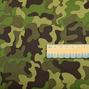 Puede incluir: Tela de camuflaje verde, marrón y beige con una regla en la parte superior. La regla es de madera y tiene un fondo blanco con marcas negras.