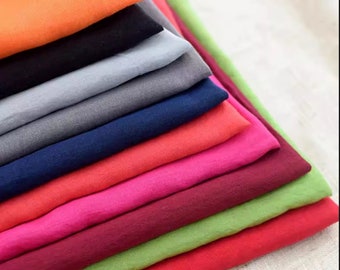 Op de afbeelding: Een stapel stofstalen in verschillende kleuren, waaronder oranje, zwart, grijs, blauw, rood, roze, bordeauxrood, groen en rood. De stofstalen zijn in een verspringende volgorde gerangschikt, met het oranje staal bovenaan.