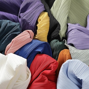 May include: A collection of pleated fabrics in various colours, including purple, green, yellow, red, blue, and white. The fabrics are arranged in a pile, with some pieces overlapping.