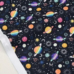 Puede incluir: Tela negra con un patrón repetido de planetas y estrellas de colores. Los planetas tienen anillos y están rodeados de varios remolinos y puntos de colores.