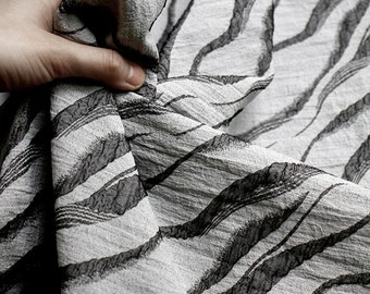 May include: A close-up of a white fabric with a black wavy pattern. The fabric is textured and has a slightly wrinkled appearance.