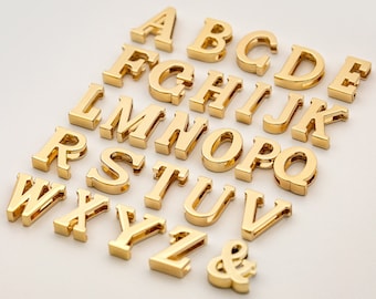 May include: Gold-colored metal alphabet letter charms, including all letters from A to Z and an ampersand symbol. The charms are arranged in a staggered pattern on a white surface.
