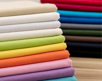 May include: A stack of folded fabric swatches in various colours, including beige, white, green, yellow, orange, pink, purple, and blue. The fabric is a smooth, woven material.