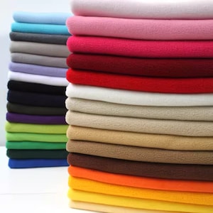 Puede incluir: Una pila de muestras de tela de polar de colores en varios tonos de azul, rosa, rojo, blanco, beige, marrón, naranja y amarillo. La tela es suave y mullida, perfecta para proyectos de manualidades y costura.