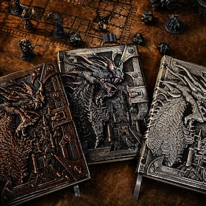 May include: Three fantasy journals with dragon designs. The journals feature embossed dragon artwork in bronze, grey, and silver tones. The scene includes miniature figurines and dice, suggesting a role-playing game theme.