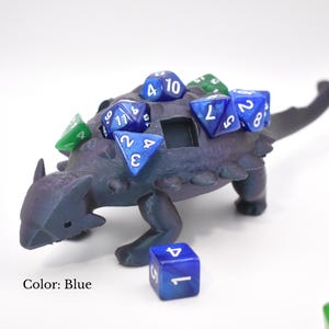 May include: A blue dinosaur-shaped dice holder with several blue and green dice on its back. The dice holder has a small opening and is surrounded by additional dice. The text "Color: Blue" is visible.