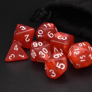 Puede incluir: Un conjunto de dados poliédricos rojos translúcidos con números blancos, cerca de una bolsa de terciopelo negro con cordón. Los dados tienen varias formas, incluyendo d4, d6, d8, d10, d12 y d20, para juegos de rol.