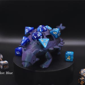 K&ouml;nnte beinhalten: Ein blauer, dinosaurierf&ouml;rmiger W&uuml;rfelhalter mit mehreren blauen W&uuml;rfeln darin und darum herum. Die W&uuml;rfel haben verschiedene Formen und Gr&ouml;&szlig;en mit wei&szlig;en und goldenen Zahlen. Der Halter steht auf einer schwarzen Oberfl&auml;che.