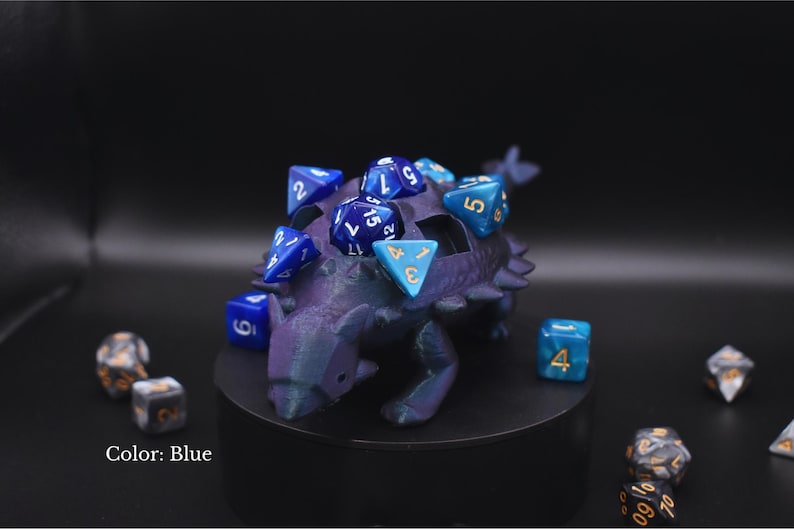 K&ouml;nnte beinhalten: Ein blauer, dinosaurierf&ouml;rmiger W&uuml;rfelhalter mit mehreren blauen W&uuml;rfeln darin und darum herum. Die W&uuml;rfel haben verschiedene Formen und Gr&ouml;&szlig;en mit wei&szlig;en und goldenen Zahlen. Der Halter steht auf einer schwarzen Oberfl&auml;che.