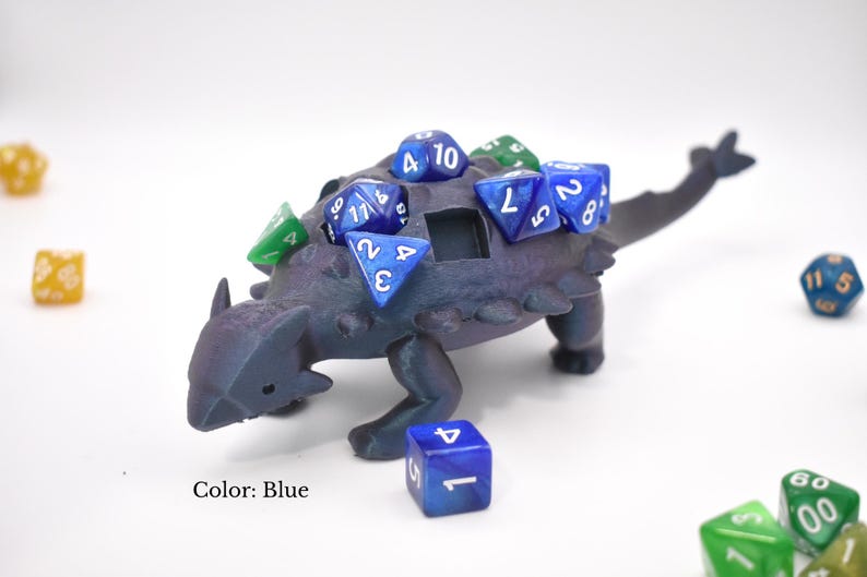 May include: A blue dinosaur-shaped dice holder with several blue and green dice on its back. The dice holder has a small opening and is surrounded by additional dice. The text "Color: Blue" is visible.
