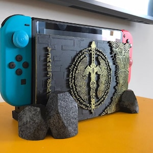 Puede incluir: Una consola Nintendo Switch negra con una funda personalizada negra y dorada que presenta una espada estilizada y un diseño circular. La consola descansa sobre un soporte hecho de cuatro piezas negras, texturizadas, con forma de roca.