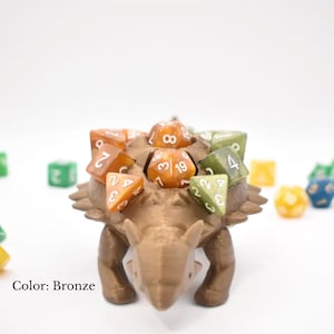 K&ouml;nnte beinhalten: Bronze-farbener W&uuml;rfelhalter in Dinosaurierform mit mehreren W&uuml;rfeln in verschiedenen Farben auf dem R&uuml;cken. Die W&uuml;rfel sind orange und gr&uuml;n. Weitere W&uuml;rfel sind um den Halter herum verstreut. Der Text "Color: Bronze" ist sichtbar.