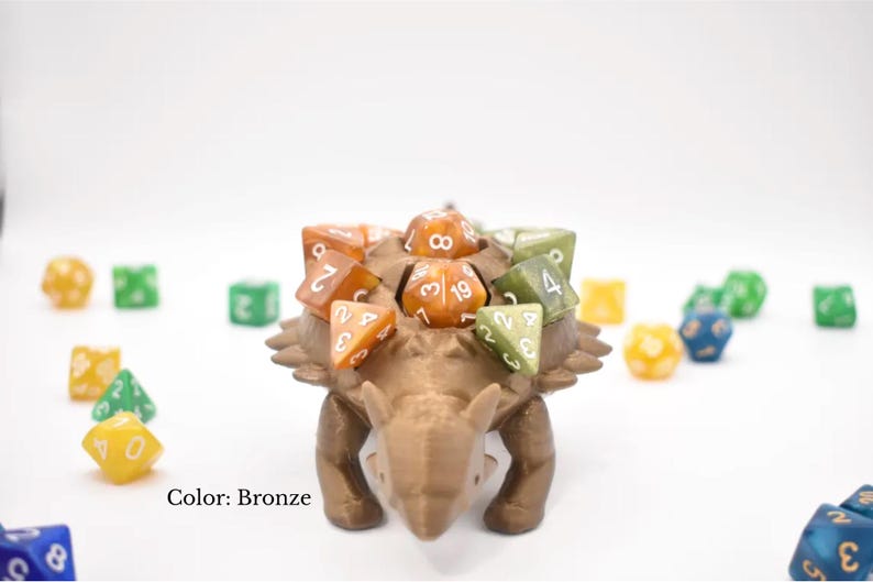 K&ouml;nnte beinhalten: Bronze-farbener W&uuml;rfelhalter in Dinosaurierform mit mehreren W&uuml;rfeln in verschiedenen Farben auf dem R&uuml;cken. Die W&uuml;rfel sind orange und gr&uuml;n. Weitere W&uuml;rfel sind um den Halter herum verstreut. Der Text "Color: Bronze" ist sichtbar.