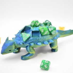 K&ouml;nnte beinhalten: Ein blau-gr&uuml;ner W&uuml;rfelhalter in Dinosaurierform mit mehreren gr&uuml;nen W&uuml;rfeln auf dem R&uuml;cken und drumherum. Der Dinosaurier hat eine strukturierte Oberfl&auml;che und einen Schlitz zur Aufnahme von W&uuml;rfeln. Die W&uuml;rfel sind in verschiedenen Gr&ouml;&szlig;en und Formen.