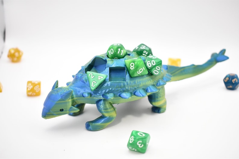 K&ouml;nnte beinhalten: Ein blau-gr&uuml;ner W&uuml;rfelhalter in Dinosaurierform mit mehreren gr&uuml;nen W&uuml;rfeln auf dem R&uuml;cken und drumherum. Der Dinosaurier hat eine strukturierte Oberfl&auml;che und einen Schlitz zur Aufnahme von W&uuml;rfeln. Die W&uuml;rfel sind in verschiedenen Gr&ouml;&szlig;en und Formen.