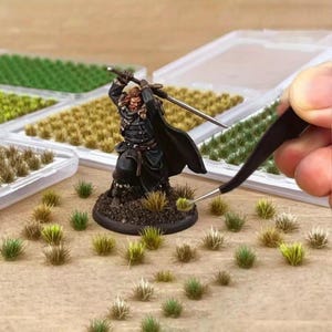 Può includere: Figurina in miniatura di un guerriero in armatura nera con una spada, circondata da ciuffi d'erba in miniatura. La scena include contenitori di erba verde e beige, e una mano che usa le pinzette per posizionare un ciuffo. Per giochi da tavolo.