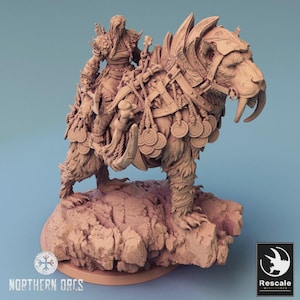 Pode incluir: Escultura em miniatura detalhada de um guerreiro montado num tigre dentes de sabre. As figuras são de cor castanha clara e montadas numa base rochosa. O tigre é adornado com elementos decorativos, e o guerreiro está equipado com armadura e armas. As palavras "Northern Orcs" estão em baixo.
