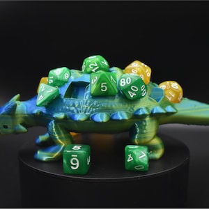 K&ouml;nnte beinhalten: Ein 3D-gedruckter, blau-gr&uuml;ner W&uuml;rfelhalter in Dinosaurierform mit mehreren gr&uuml;nen und gelben W&uuml;rfeln auf dem R&uuml;cken. Zwei gr&uuml;ne W&uuml;rfel befinden sich vor dem Halter. Der Dinosaurier hat ein detailliertes, strukturiertes Design.