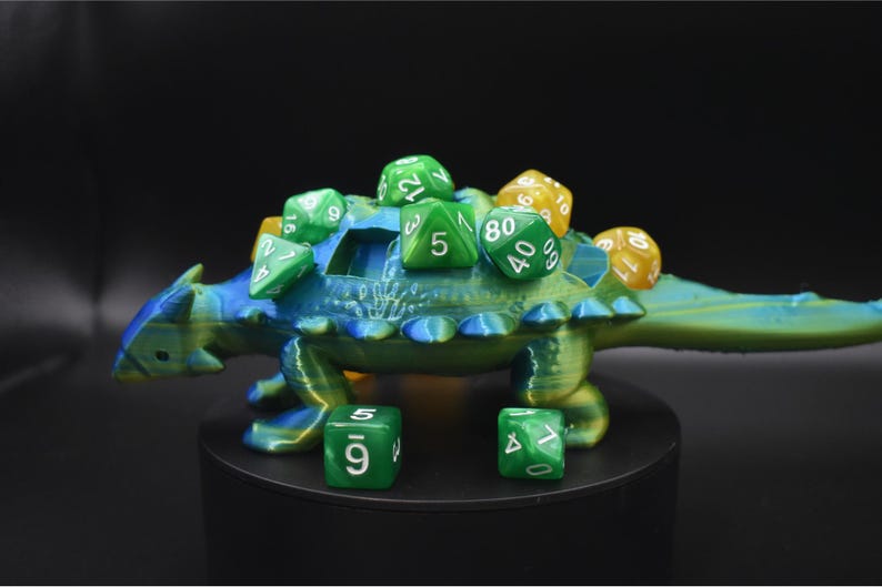 K&ouml;nnte beinhalten: Ein 3D-gedruckter, blau-gr&uuml;ner W&uuml;rfelhalter in Dinosaurierform mit mehreren gr&uuml;nen und gelben W&uuml;rfeln auf dem R&uuml;cken. Zwei gr&uuml;ne W&uuml;rfel befinden sich vor dem Halter. Der Dinosaurier hat ein detailliertes, strukturiertes Design.