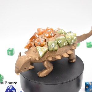 K&ouml;nnte beinhalten: Bronzefarbener Dinosaurier-W&uuml;rfelhalter mit verschiedenen bunten W&uuml;rfeln. Der Dinosaurier hat einen strukturierten R&uuml;cken und ist von anderen W&uuml;rfeln in verschiedenen Farben umgeben. Der Text "Color: Bronze" ist sichtbar.