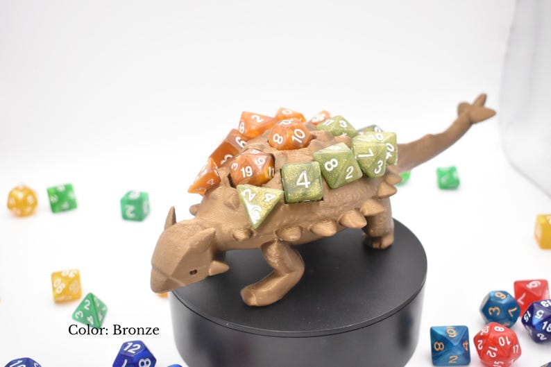 K&ouml;nnte beinhalten: Bronzefarbener Dinosaurier-W&uuml;rfelhalter mit verschiedenen bunten W&uuml;rfeln. Der Dinosaurier hat einen strukturierten R&uuml;cken und ist von anderen W&uuml;rfeln in verschiedenen Farben umgeben. Der Text "Color: Bronze" ist sichtbar.