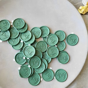May include: A pile of green wax seals with a cursive letter "B" on each seal. The seals are on a light gray plate.