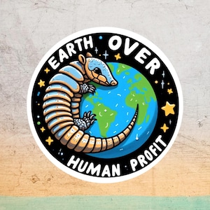 May include: A round sticker featuring an armadillo wrapped around a blue and green globe. The text "EARTH OVER HUMAN PROFIT" is written around the edge in white. The armadillo is blue and tan with a smiling face and the background is black with yellow stars.
