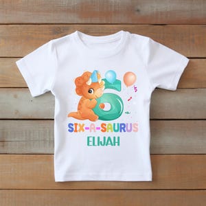 May include: White t-shirt with a colorful graphic featuring a cartoon dinosaur wearing a party hat, the number 6, and balloons. Text on the shirt reads "SIX-A-SAURUS ELIJAH". The design is in shades of orange, teal, blue, and pink.