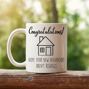 May include: White ceramic mug with a black house graphic and the text "Congratulations! HOPE YOUR NEW NEIGHBOURS AREN'T ASSHOLES". The mug has a curved handle and is set against a blurred green background.