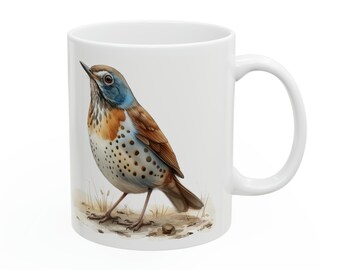 Puede incluir: Taza de cerámica blanca con una ilustración de acuarela de un pájaro. El pájaro tiene plumaje marrón, naranja y azul y un pecho moteado. La taza tiene un asa curva y está sobre un fondo blanco.