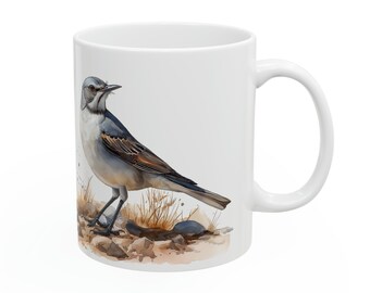 Puede incluir: Taza de cerámica blanca con una ilustración de acuarela de un pájaro con plumas grises, marrones y blancas. El pájaro está posado sobre una superficie rocosa con hierba. La taza tiene un asa curva.