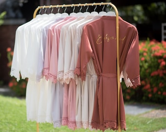 May include: A set of eight satin robes with lace trim, hanging on a gold rack. The robes are in various shades of pink, white, and beige. One robe has the word "Bridemaid" embroidered on the back in gold lettering.