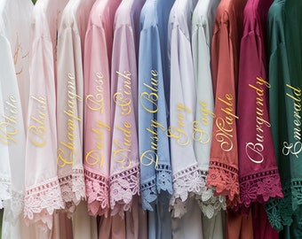 May include: A row of 10 satin robes in various colours, including white, blush, champagne, dusty rose, nude pink, dusty blue, grey, sage, maple, burgundy, and emerald. Each robe has a lace trim.