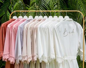 May include: A set of 10 satin robes with lace trim, hanging on a gold rack. One robe has the word "Bride" embroidered on the back in gold lettering.