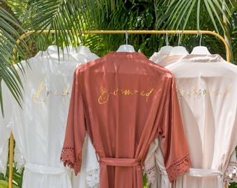 May include: Three satin robes with lace trim, hanging on a gold rack. The robes are white, rust, and blush pink. Each robe has the word "Bridesmaid" embroidered in gold on the back.