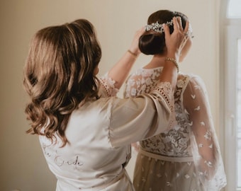 Puede incluir: Una mujer con una bata de satén blanca con el texto "The Bride" bordado en la espalda está ayudando a otra mujer a ponerse una corona floral. La mujer que lleva la corona lleva un vestido de encaje blanco.