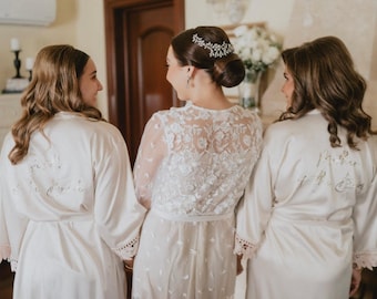 Puede incluir: Tres mujeres con batas de satén blanco con ribetes de encaje y texto bordado. La novia está en el centro con un vestido de encaje blanco y una tiara.