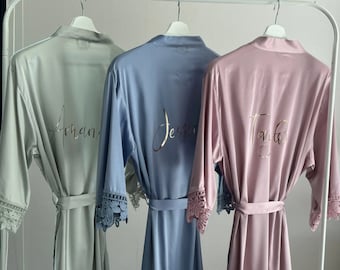 May include: Three satin robes in different colors, light green, light blue, and light pink, with lace trim on the sleeves. Each robe has a name written on the back in gold lettering: "Amanda", "Jessica", and "Taylor".