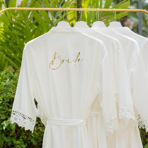 May include: Four white robes with lace trim hang on a gold rack. The robe on the left has the word "Bride" written in gold script on the back.