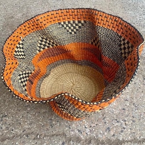 May include: A woven basket with a unique, wavy rim. The basket features a blend of orange, black, and natural beige tones. The design includes geometric patterns and a circular base, showcasing a handcrafted aesthetic.