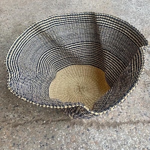 Puede incluir: Cesta grande tejida con base redonda y laterales acampanados. La cesta presenta rayas horizontales alternas en negro y beige natural. El interior es de color beige natural. La cesta está sobre una superficie de hormigón.
