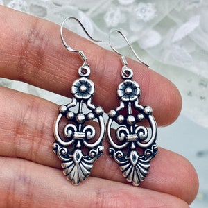 May include: A pair of silver dangle earrings with a floral design. The earrings have a delicate, intricate design with a flower at the top and a series of small beads and swirls.