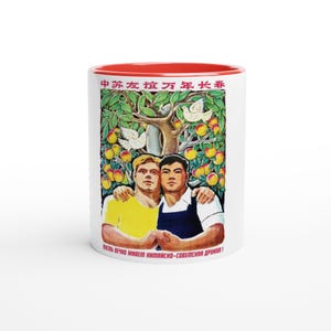 Puede incluir: Taza de cerámica blanca con interior y borde rojos. La taza presenta una ilustración vintage de dos hombres abrazándose bajo un árbol con palomas y fruta. Texto chino y ruso visible.