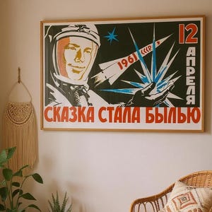 May include: A framed vintage poster with a cosmonaut illustration, a rocket, and a burst of blue and white light. The poster has red and white Cyrillic text and the date "1961". The poster is in a wooden frame, ideal for a retro aesthetic.