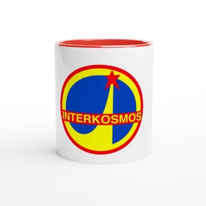 Op de afbeelding: Een witte keramische mok met een rode binnenrand. De voorzijde is voorzien van een rond logo met een gele rand, een blauwe achtergrond, een gele gestileerde raketvorm, een rode ster en de tekst INTERKOSMOS in gele letters in het midden.