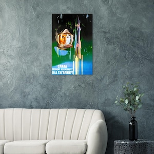 Puede incluir: Un póster rectangular vertical que presenta el retrato de un cosmonauta con casco blanco sobre un fondo espacial oscuro con un cohete despegando. El texto en cirílico blanco dice SLAVA PERVOMU KOSMONAVTU YU.A. GAGARINU.